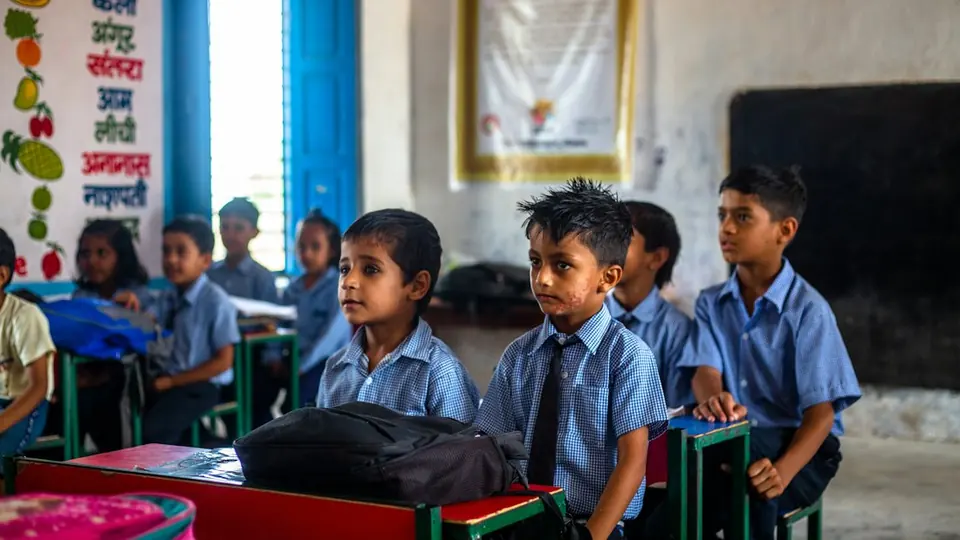 Children in blue uniforms sitting at desks in a classroom, with a blackboard behind (stock photo).