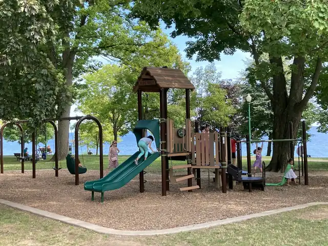 A wooden climbing frame with a green slide and swings in a playground shaded by tall trees (stock photo).