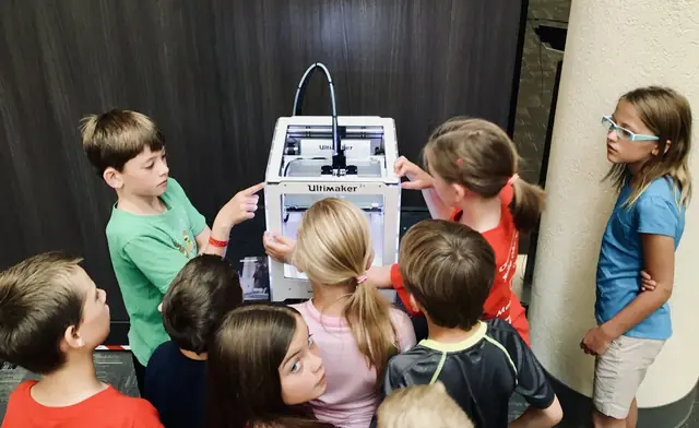 A group of children crowding around a 3D printer and watching it work (stock photo).